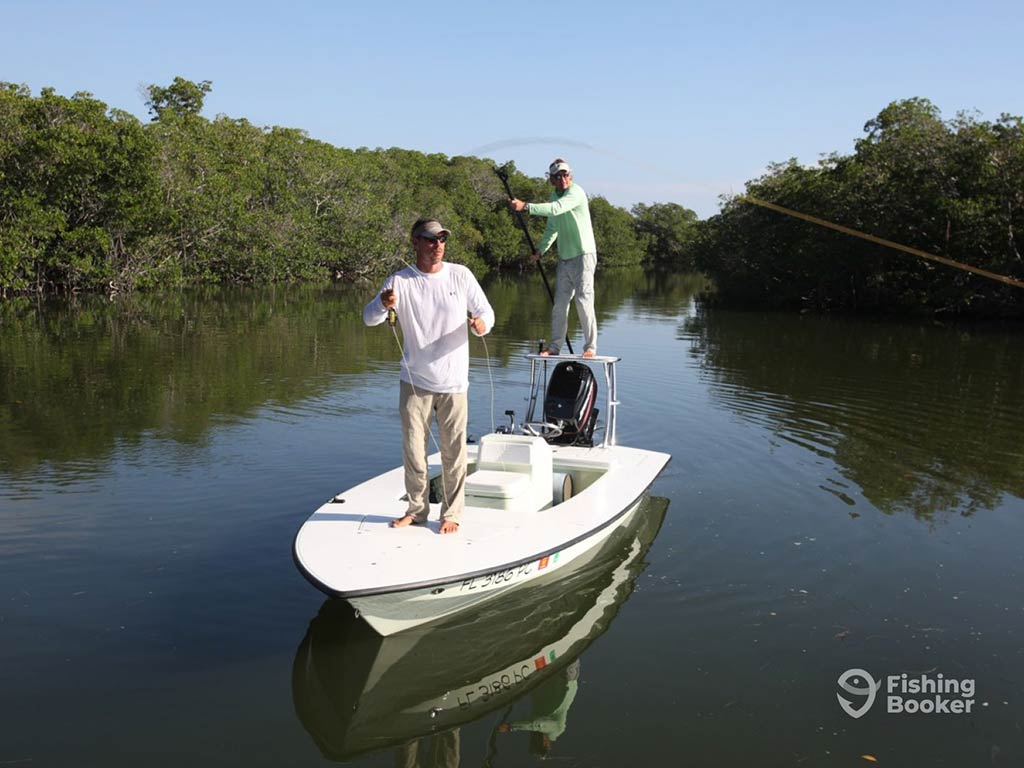 Two anglers on a skiff fishing boat in the calm backcountry waters of Florida, with mangrove-lined shores either side of them, as one man is polling the boat from the rear and another is preparing to cast a fly fishing line