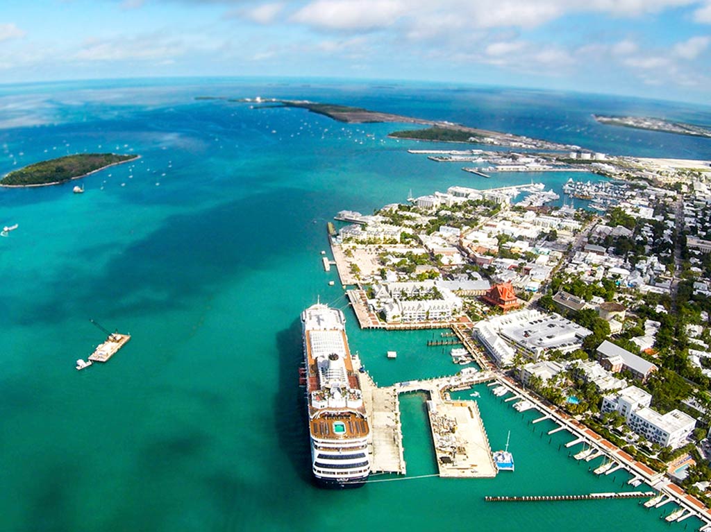 An aerial view of Key West, with a cruise ship docked in the foreground of the image, the town on the right, and numerous islands dotted in the crystal clear waters all the way to the horizon