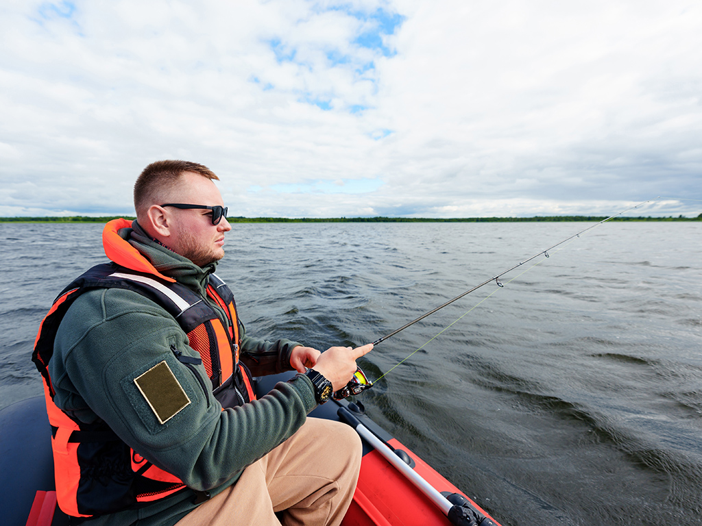 A fisherman in a red life jacket and sunglasses, sitting in a red boat with a fishing rod, while fishing on a lake in summer