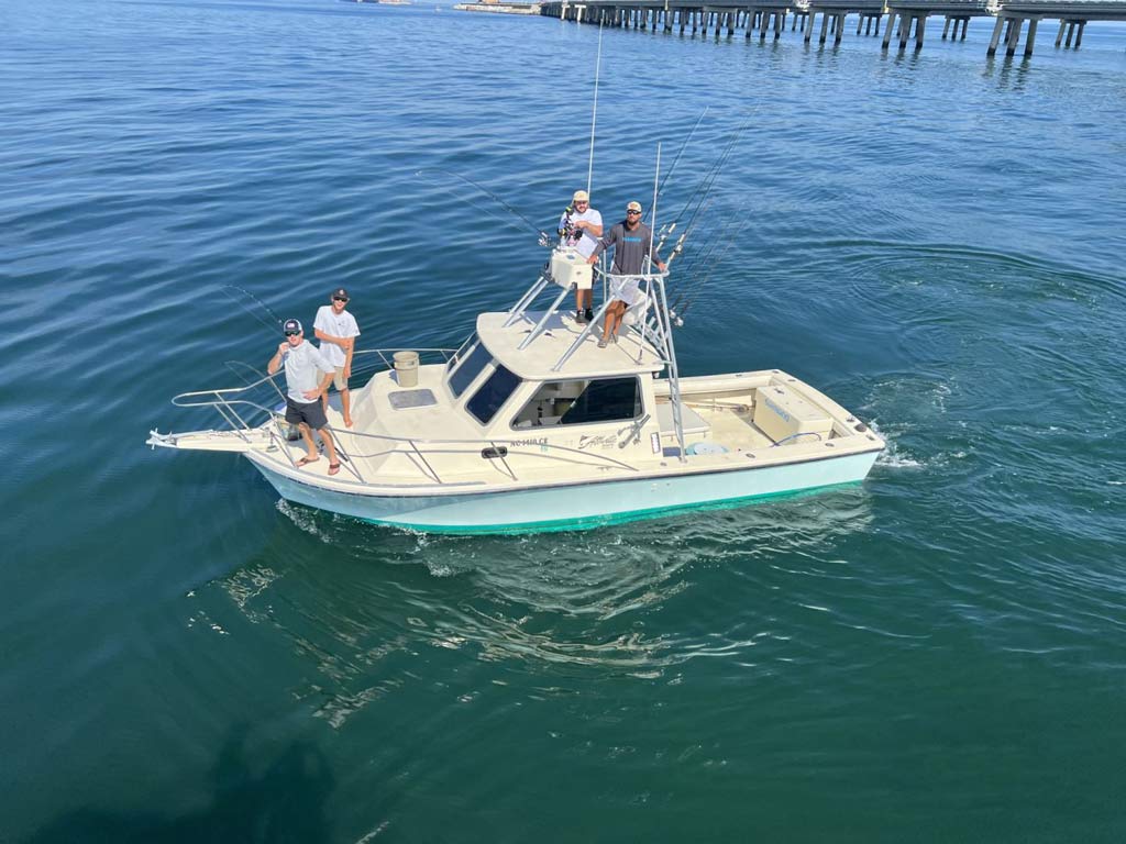 An aerial shot of a charter fishing boat with 4 people on it, 2 are standing on the flybridge while the other two are at the bow, the group is getting ready for a summer fishing adventure.