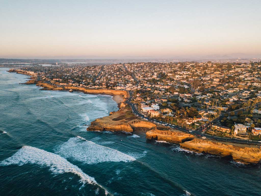 An aerial photo of San Diego's shores, with waves crashing against the city's rocky cliffs and numerous buildings and houses stretching towards the horizon.