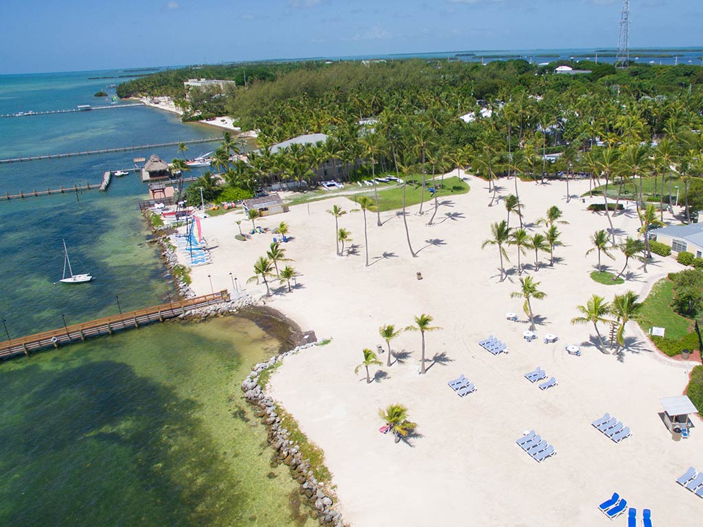 An aerial view of a beach in Islamorada, FL in summer, with sun loungers visible in the foreground and a fishing pier sticking out into the water in the middle of the image.