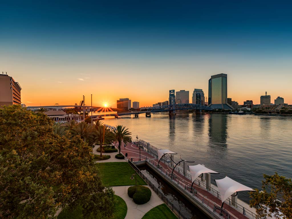 A view of the Jacksonville Riverwalk at sunset, with the St. Johns River visible to the right of the photo and numerous buildings in the distance.