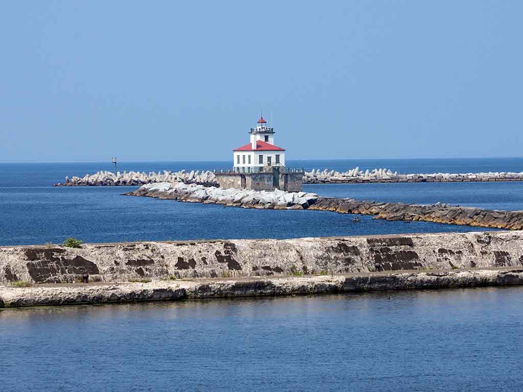 A view towards Oswego Lighthouse and Lake Ontario, with a jetty visible in the foreground on a clear day, with calm waters in the distance.