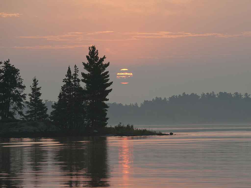 A view across the calm waters of Lake of the Woods at sunset, with the sun visible behind some low-lying clouds and trees in the distance.