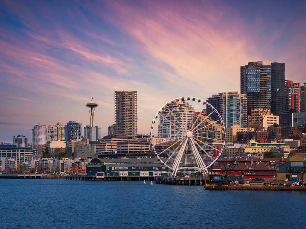 A sunset view of Seattle’s skyline and several iconic landmarks posing against the blue water in front of them and purple-hued skies behind them.