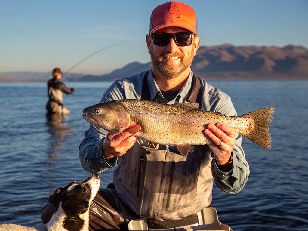 A man in a red hat and sunglasses crouches and poses with a Cutthroat Trout caught in Nevada on a sunny day, with another man out of focus in the distance casting a line
