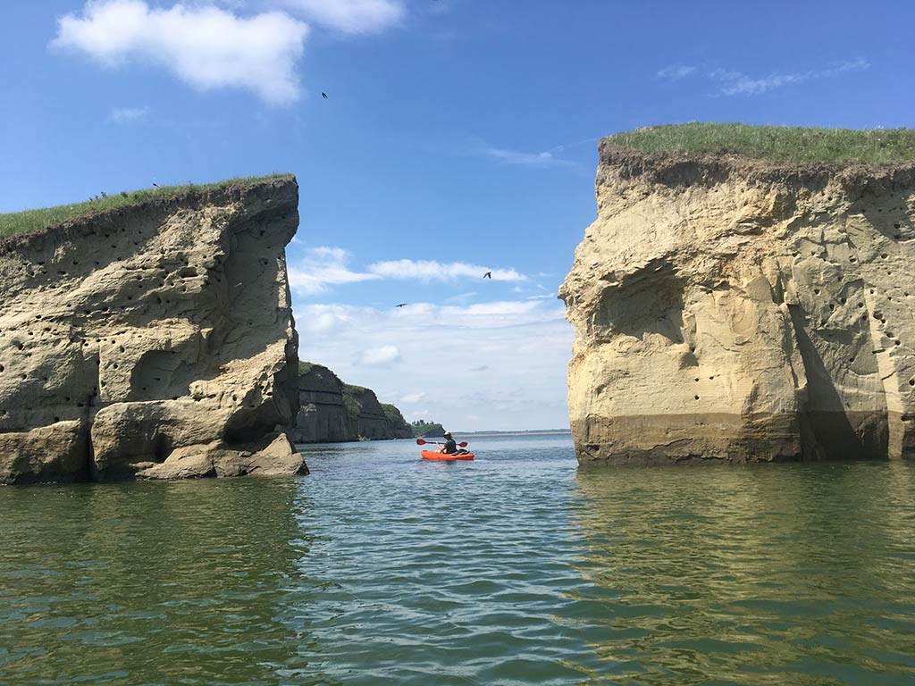 A view looking towards a gorge between two ricks in Government Bay, Lake Sakakawea, North Dakota, on a clear day, with a lone kayaker paddling through the gap and birds flying above on a clear day