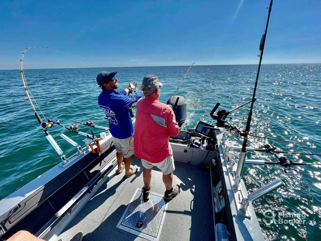 A photo featuring two anglers standing on a charter fishing boat and facing the ocean while one of them is fishing on a sunny summer day near the Fourth of July