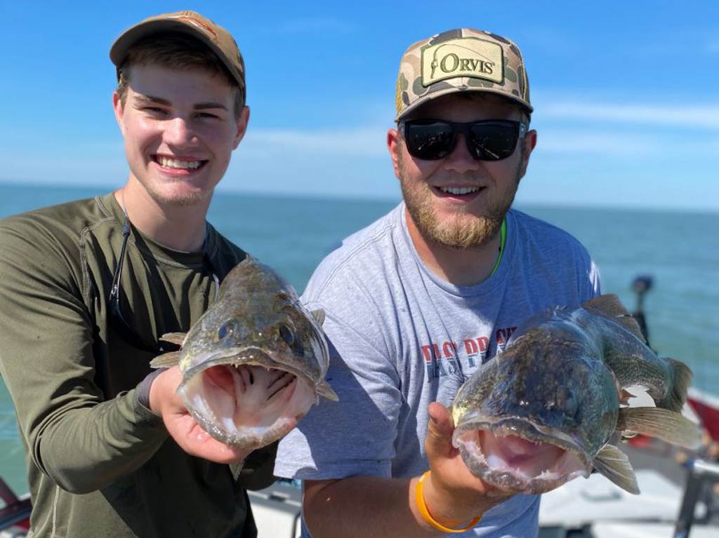 A photo featuring two proud and happy anglers standing on a charter fishing boat and posing with a Walleye each after their Fourth of July fishing trip, with their Walleyes in their hands, facing towards the camera mouths open