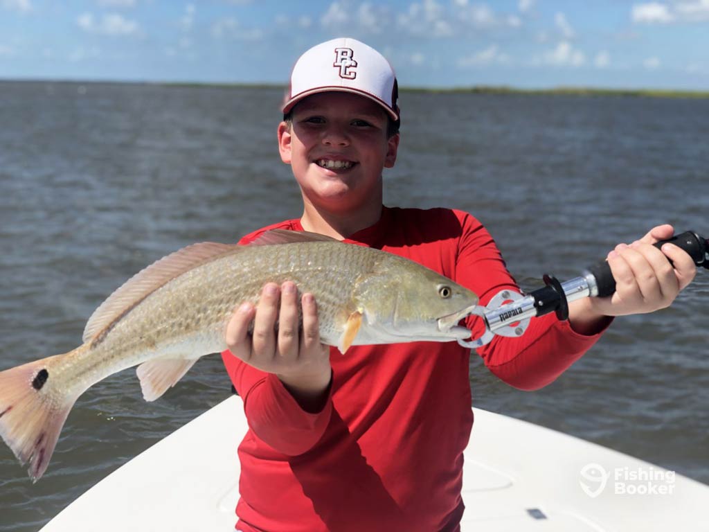 A photo featuring a happy kid wearing a baseball cap, posing with a Redfish caught on a Fourth of July fishing trip out of Port Arthur in Texas
