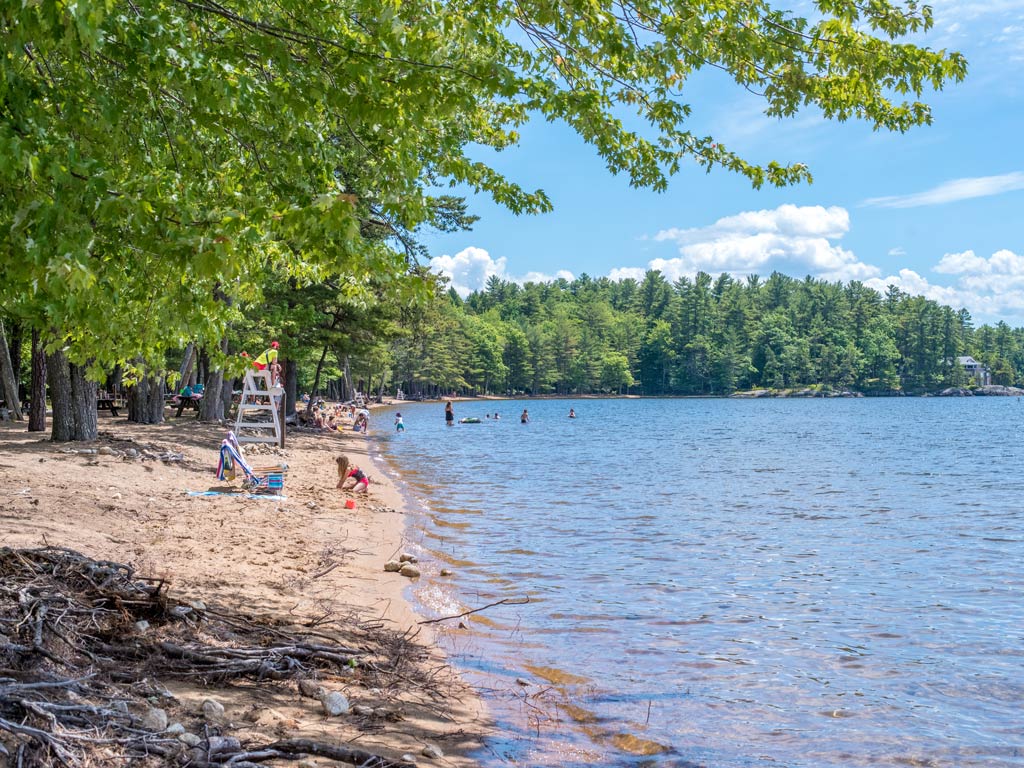 A photo featuring beachgoers on a sandy shoreline of Sebago Lake in Maine, with swimmers in the water refreshing during summer 