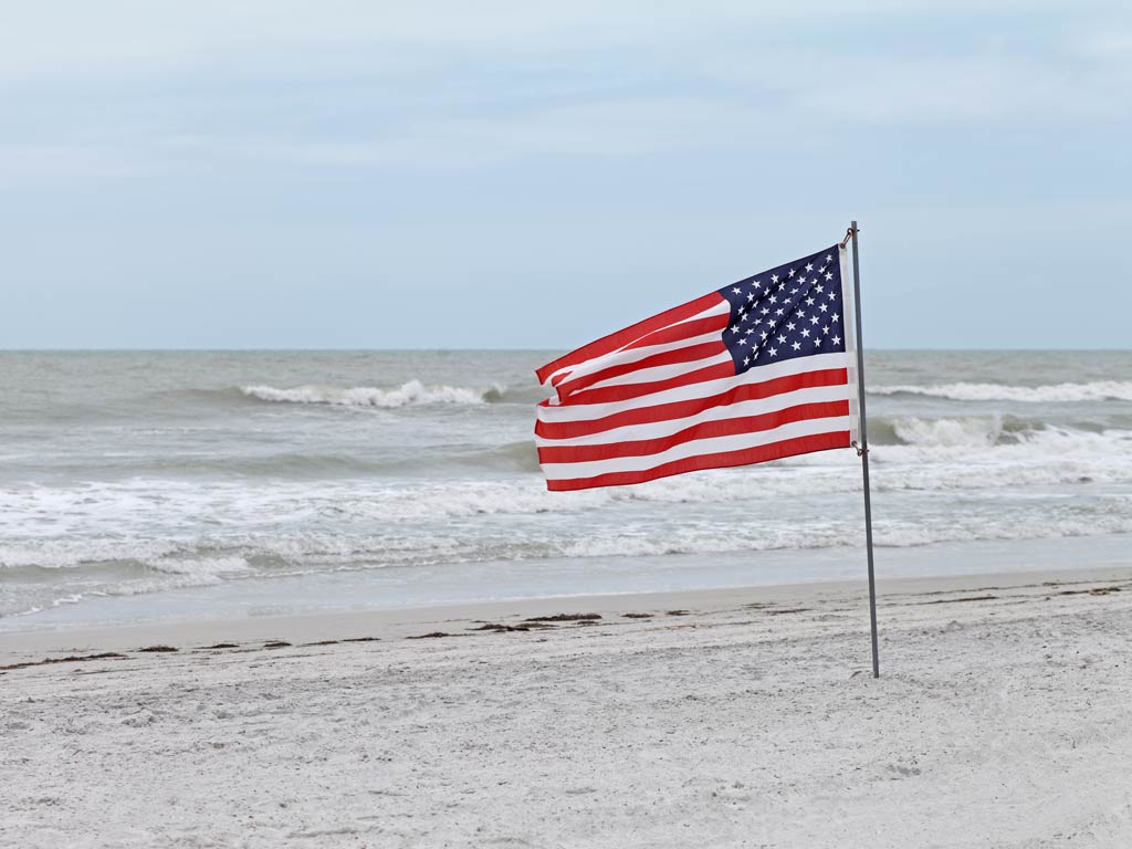 A photo features American flag proudly blowing in the wind placed in the sand at a beach next to the ocean