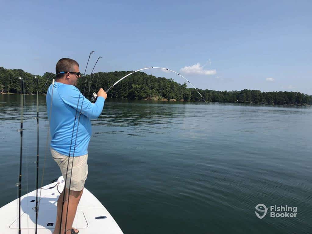 An angler in a blue shirt and cream shorts casts a line into the calm waters of Lake Lanier on a clear day from the front of a fishing boat